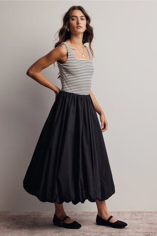 A woman modeling a sleeveless midi dress with a black and white horizontally striped bodice and a voluminous black skirt, paired with black flats, against a neutral studio background.