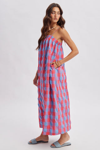 A woman modeling a sleeveless midi dress with a pink and light blue gingham pattern, paired with gray slide sandals, against a plain light gray studio background.