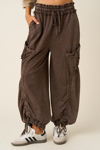 A person wearing loose brown cargo jogger pants with drawstrings, side cargo pockets, and elastic cuffs, paired with white sneakers, against a plain light beige studio background.