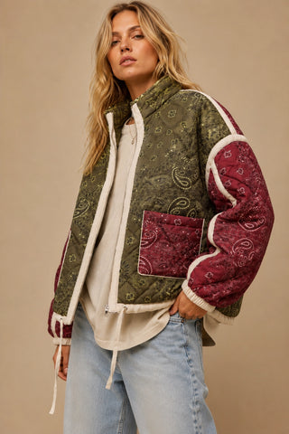 A woman wearing a patchwork quilted jacket with an olive green paisley pattern, burgundy sleeves and pocket, and cream sherpa lining, paired with a cream top and light blue jeans against a solid beige background.