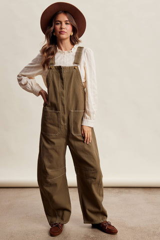 A woman modeling wide-leg olive brown utility overalls over a white long-sleeve blouse, wearing a brown wide-brim hat and brown loafers, against a neutral beige studio background.