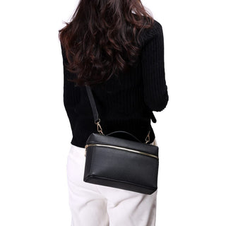 A woman shown from the back wearing a black ribbed sweater and white pants, carrying a black pebbled leather top-handle handbag with a gold zipper and strap over her shoulder.