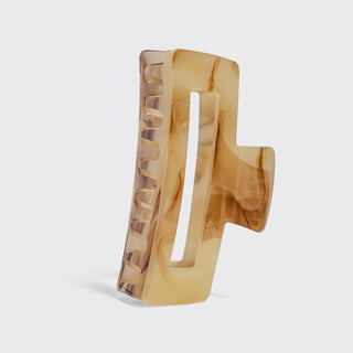 A single large hair claw clip in a translucent amber-brown acetate with natural marbled swirls, shown standing upright on a light gray background.