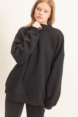 A woman wearing an oversized black sweatshirt with a mock neck and long sleeves, paired with dark gray jeans, photographed against a plain off-white studio background.