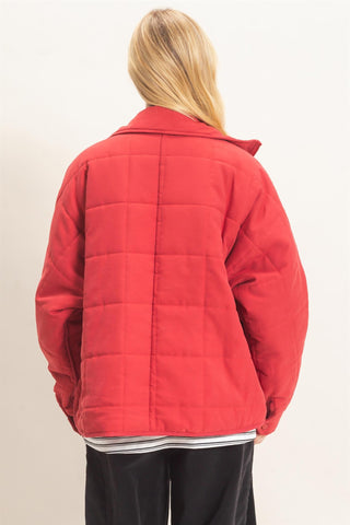 A woman viewed from behind wearing a bright red quilted jacket, black pants, and a striped shirt against a plain off-white background.