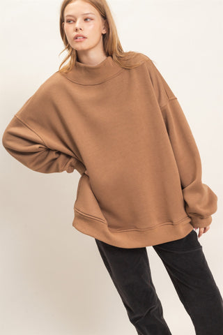 A woman wearing an oversized camel brown sweatshirt and black pants, photographed against a plain light beige studio background.