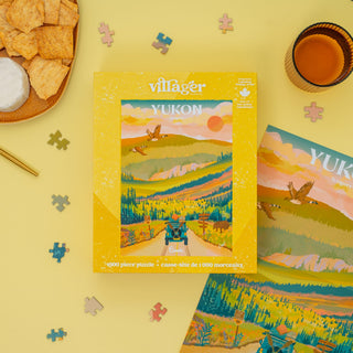 Villager Yukon puzzle box on a yellow surface with puzzle pieces and snacks.