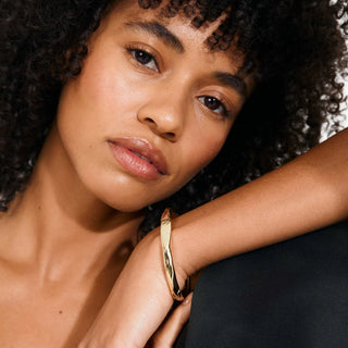A close-up lifestyle shot of a model wearing a polished gold hinged bangle bracelet on her wrist, resting her arm on a dark surface.