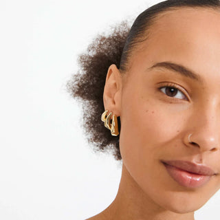 A close-up of a model wearing a pair of gold triple-hoop earrings against a clean white background.