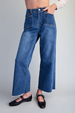 A person wearing high-waisted, wide-leg cropped blue denim jeans with a raw hem, styled with black Mary Jane flats against a light gray studio background.
