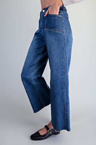 A person wearing high-waisted, wide-leg blue denim jeans with a frayed hem, paired with black Mary Jane flats, shown from the waist down against a light gray studio background.