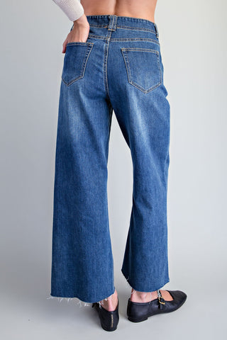 A person shown from the back wearing blue wide-leg cropped jeans with a frayed hem, paired with black flats, against a light gray studio background.