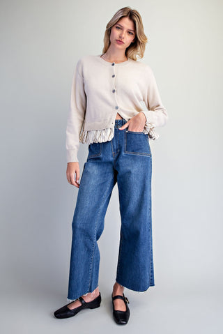 A woman modeling a cream-colored cardigan with fringe trim and wide-leg blue jeans against a light gray studio background.