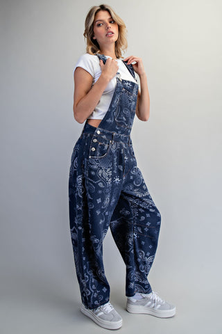 A woman modeling dark blue denim overalls with a white paisley bandana print, styled with a white crop top and white sneakers against a light gray studio background.
