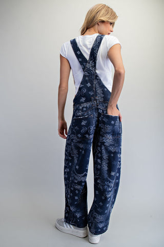 A model shown from the back wearing dark blue bandana-print denim overalls over a white t-shirt and white sneakers, set against a plain light gray studio background.