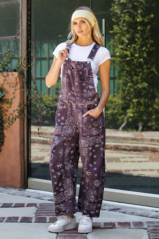A woman wearing a pair of loose-fitting purple paisley bandana print overalls over a white t-shirt and white sneakers, standing outdoors in front of a glass door.