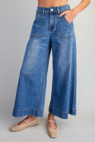 A model wearing high-waisted, wide-leg blue denim jeans with front patch pockets and rolled hems, paired with woven beige flat shoes, against a neutral gray studio background.