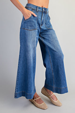 A person wearing high-waisted, wide-leg blue denim jeans with front patch pockets and rolled cuffs, paired with beige woven flat shoes against a light gray studio background.