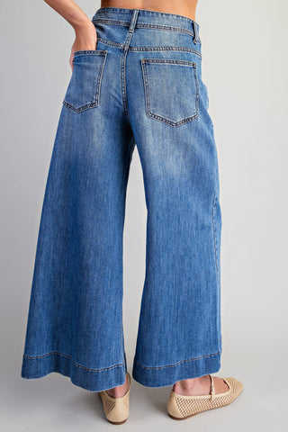 A person wearing blue wide-leg denim jeans, shown from the back, paired with beige flat shoes against a light gray studio background.