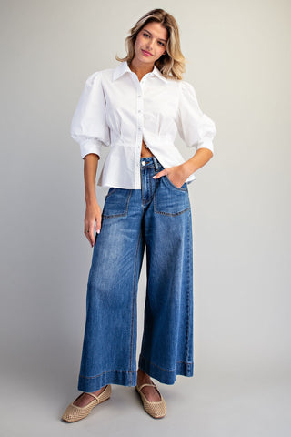 A woman modeling a white long-sleeve blouse with puffed sleeves and blue wide-leg denim jeans, paired with beige flat shoes, against a light gray studio background.