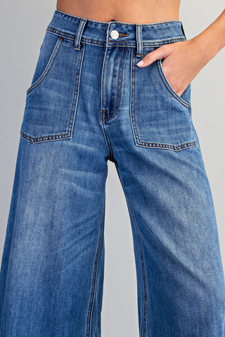 Close-up of a person wearing high-waisted wide-leg blue denim jeans with front patch pockets and a button closure, shown against a light gray studio background.