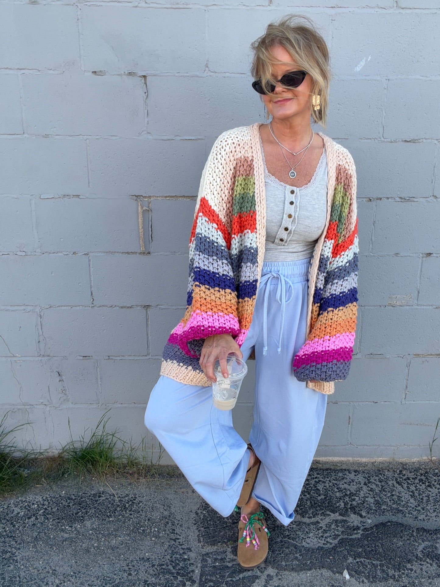 Woman wearing a colorful striped cardigan over a white dress against a gray wall.