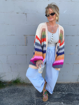 Woman wearing a colorful striped cardigan over a white dress against a gray wall.