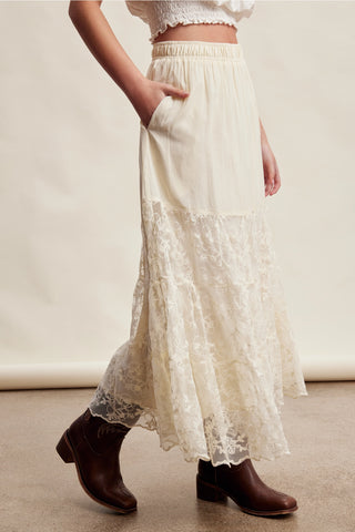 A model wearing a cream-colored, high-waisted maxi skirt with a lace hem and brown leather ankle boots, standing against a neutral cream backdrop.