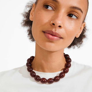 A close-up of a model wearing a dark reddish-brown beaded necklace with polished round beads, styled with a white crew-neck top against a clean white background.