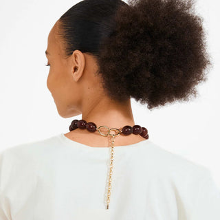 A woman shown from the back wearing a dark brown beaded necklace with a gold-tone ring and dangling chain, paired with a white top against a light gray background.