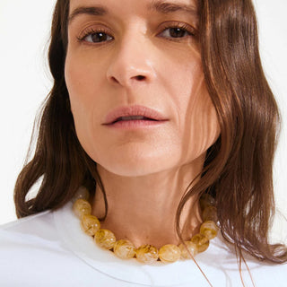A close-up of a woman wearing a chunky amber-yellow beaded necklace with a white top against a plain light background.
