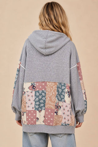 A woman shown from the back wearing a heather gray hoodie with a large patchwork panel on the back and sleeves in dusty pink, beige, and blue floral prints, paired with light blue jeans against a solid beige background.