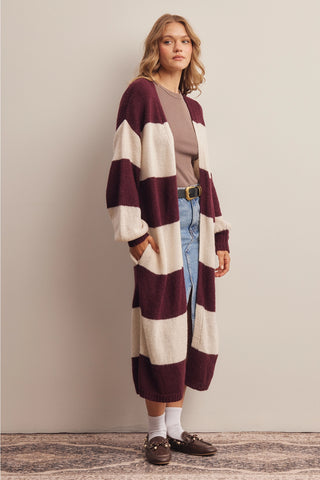 A woman models a long, open-front knit cardigan with wide horizontal stripes in deep burgundy and cream. She is wearing a taupe top, blue jeans, white socks, and brown loafers, standing against a neutral beige wall.