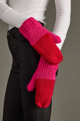 Pair of pink and red knitted mittens held by a person wearing a white sweater and black pants.