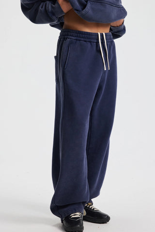 A person wearing navy blue sweatpants with white drawstrings, paired with black loafers, shown from the waist down against a light gray studio background.