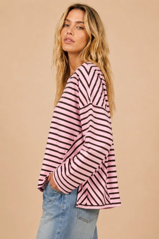 A woman wearing a pink and dark red striped long-sleeve top with blue jeans, standing against a solid beige background.