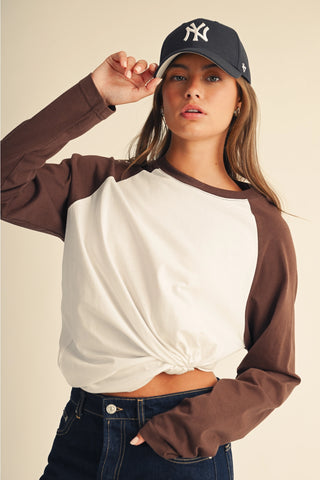 A woman wearing a brown and off-white raglan long-sleeve crop top, dark blue jeans, and a navy New York Yankees baseball cap, posing against a solid beige background.