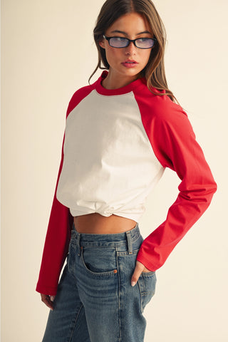 A woman wearing a long-sleeve raglan crop top with a white body and red sleeves, paired with blue jeans, photographed against a plain beige studio background.