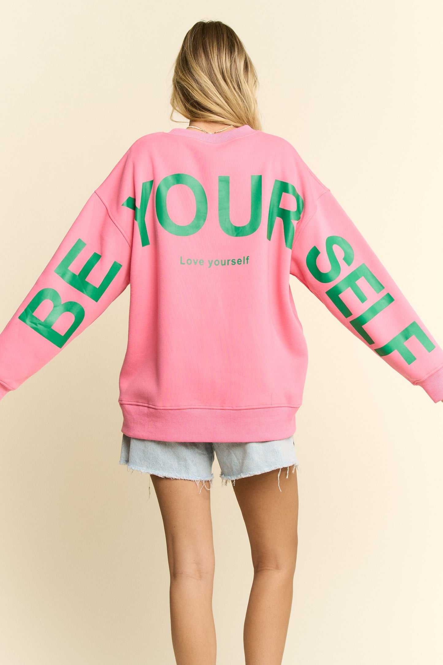 Person wearing a pink sweatshirt with 'BE YOURSELF' text in green on a beige background