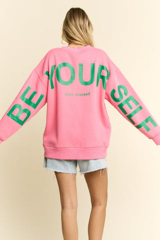 Person wearing a pink sweatshirt with 'BE YOURSELF' text in green on a beige background