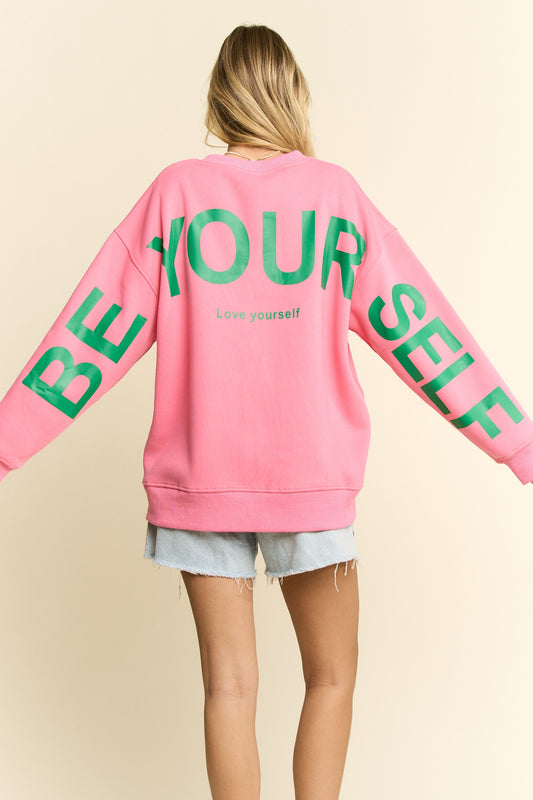 Person wearing a pink sweatshirt with 'BE YOURSELF' text in green on a beige background