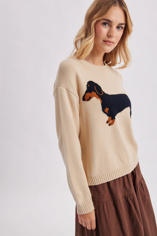 A woman wearing a cream-colored knit sweater with a black and brown dachshund graphic on the chest, paired with a brown skirt, against a light gray studio background.