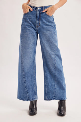 A person wearing blue wide-leg denim jeans with hands in the pockets, paired with a white top and black ankle boots, against a plain light gray studio background.