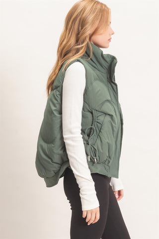 A woman shown from the side wearing an olive green puffer vest over a white long-sleeve top and black pants, photographed against a plain light background.