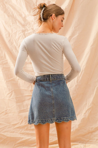 Woman wearing a beige long-sleeve top and denim skirt against a beige background