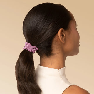 A woman shown from the back and side profile wearing a textured, ruffled light purple scrunchie that holds her dark brown hair in a low ponytail, set against a solid beige background.