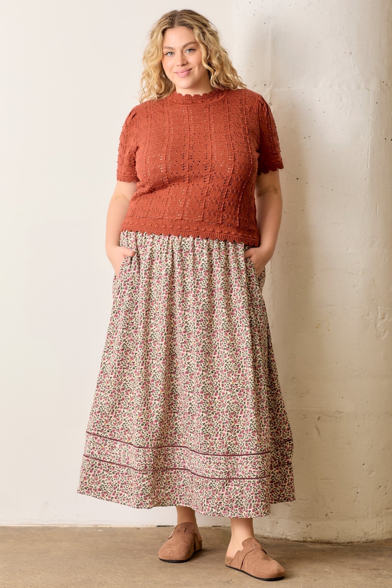 Woman wearing a rust-colored top and patterned skirt against a plain background