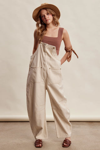 Woman wearing beige overalls and a brown hat against a neutral background