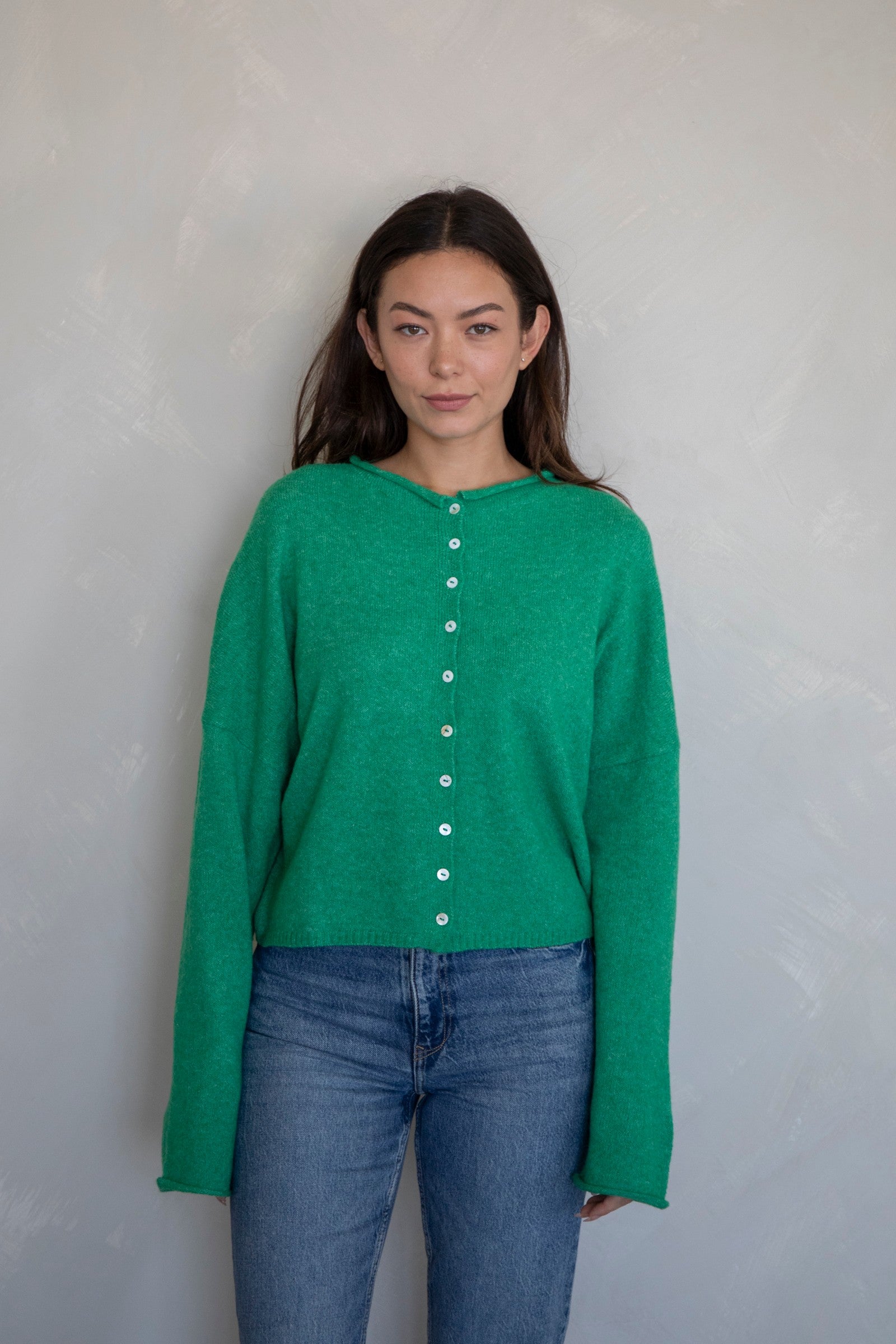 Woman wearing a green long-sleeve shirt with buttons and blue jeans against a plain background