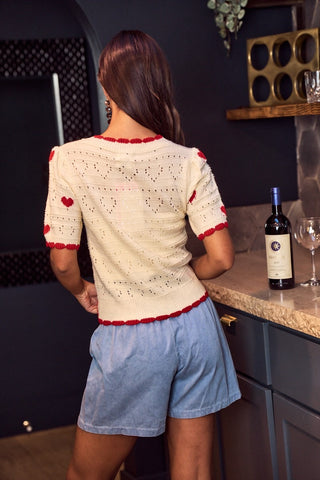 Woman in a white crochet top with red accents and blue shorts standing in a kitchen.